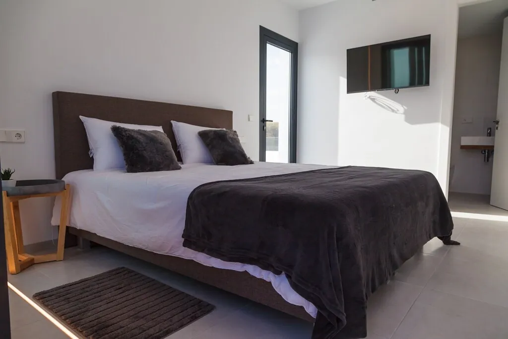 Ein Schlafzimmer mit dunkler Tagesdecke, einem Teppich neben dem Bett und einem Fernseher an der Wand.
