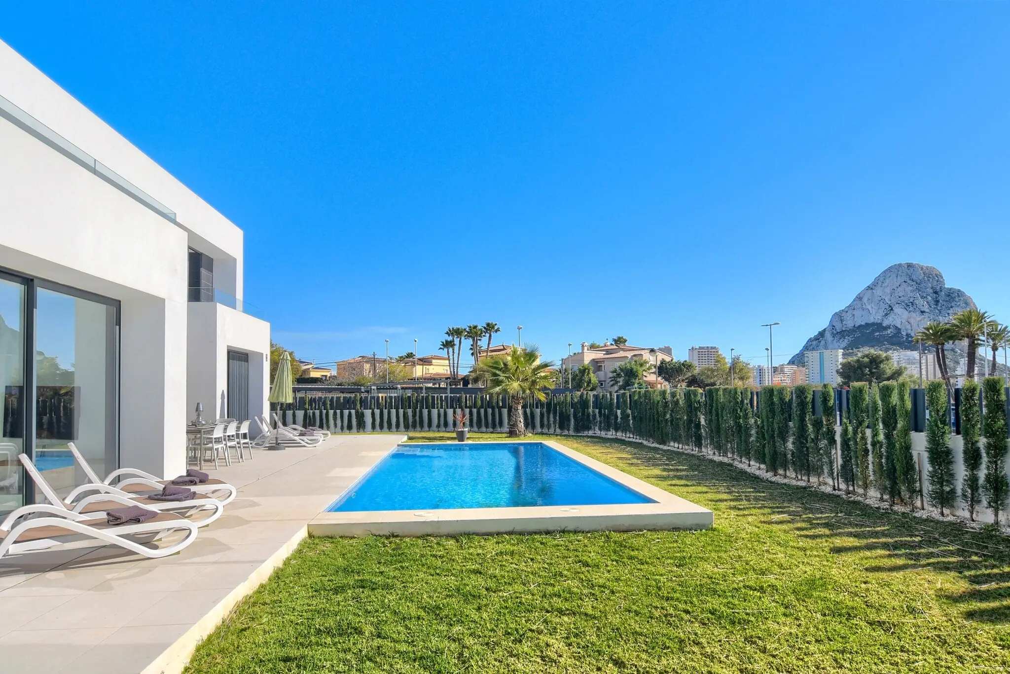 Villa Clem von der Gartenseite: ein weißes zweigeschossiges Haus, der Pool im gemähten Rasen, Liegen entlang der Kante und rechts der Felsen Peñón de Ifach.