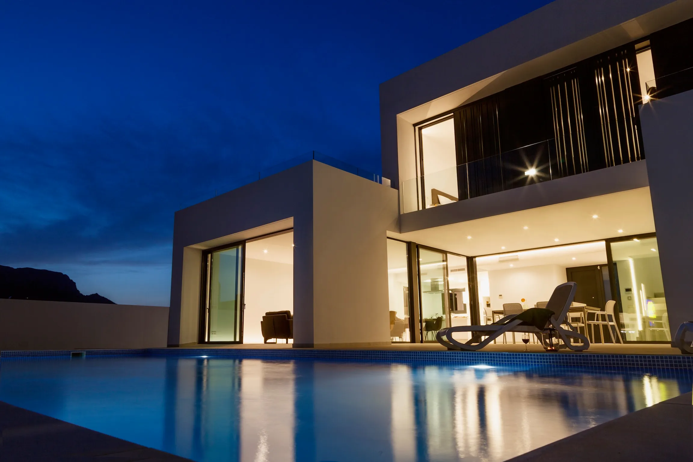 Dasselbe Haus nach Sonnenuntergang, alle Räume beleuchtet und der Pool blau leuchtend.