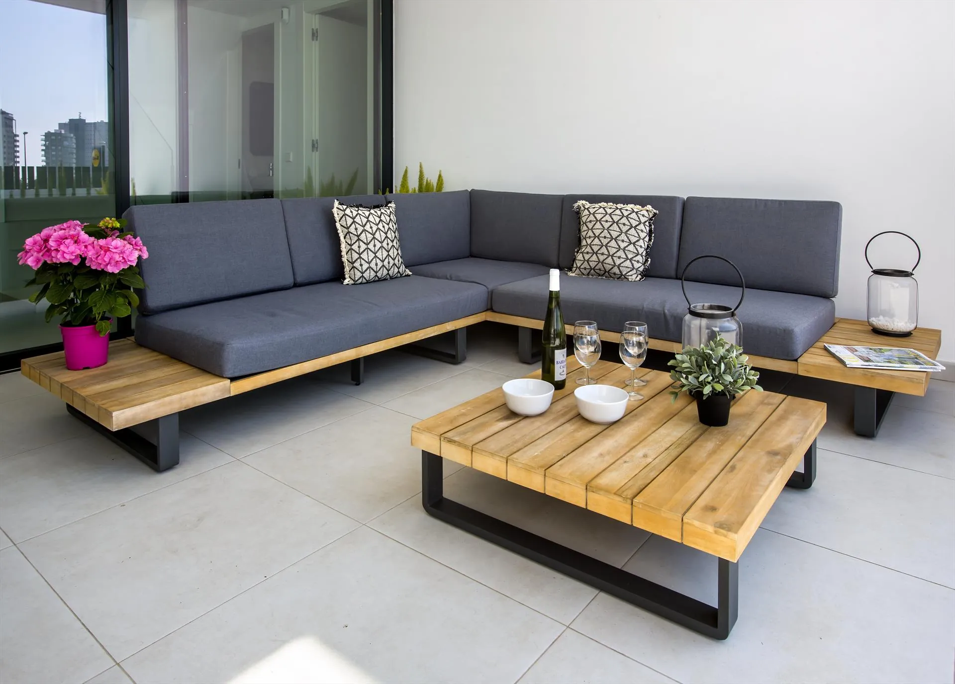 Die überdachte Terrasse mit dunkelgrauem Ecksofa um zwei niedrige Holztische, Blumen und Laternen.